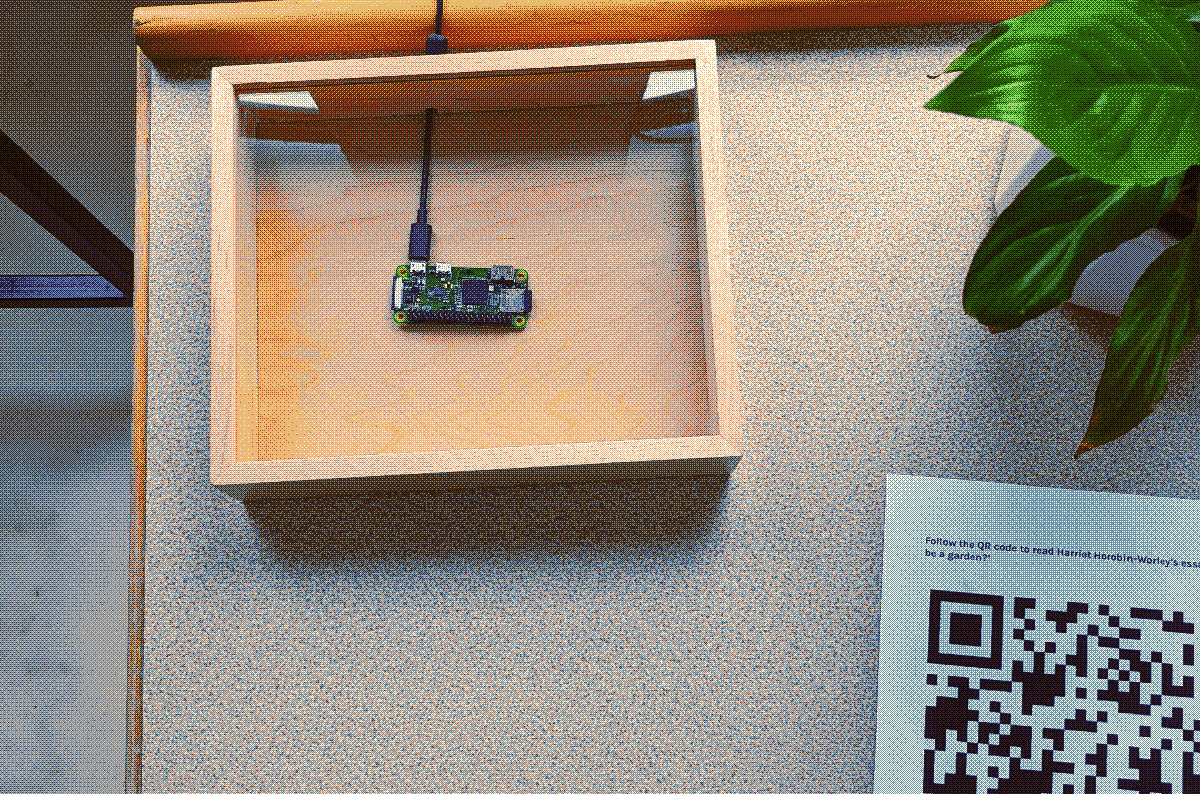 a top-down view of a raspberry pi in a handcrafted box, next to a plant and a bit of a QR code on a piece of paper, on a table